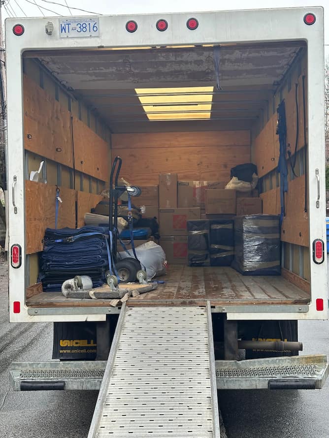 The open back of a Unicell moving truck is filled with cardboard boxes, blue moving blankets, and a hand truck with a ramp extended to the ground.